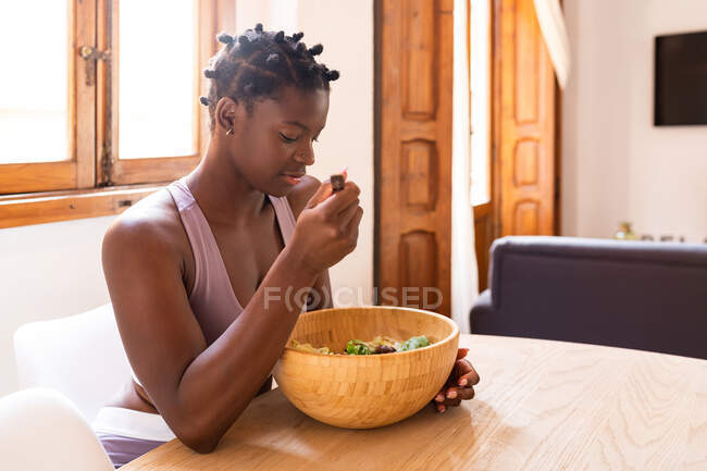 Vista lateral de la hembra afroamericana comiendo ensalada de verduras frescas mientras está sentada en la mesa y almorzando saludable en casa - foto de stock