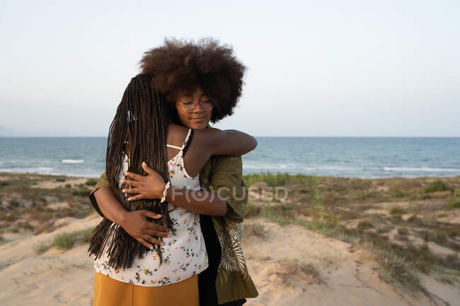 Feliz joven afroamericana hembra mejores amigos abrazándose mientras pasan las vacaciones de verano juntos en la orilla del mar - foto de stock
