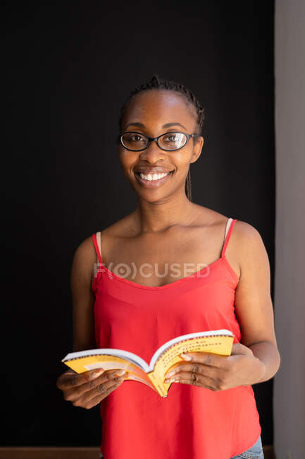 Donna afroamericana con acconciatura intrecciata e in bicchieri lettura interessante storia in libro su sfondo nero — Foto stock