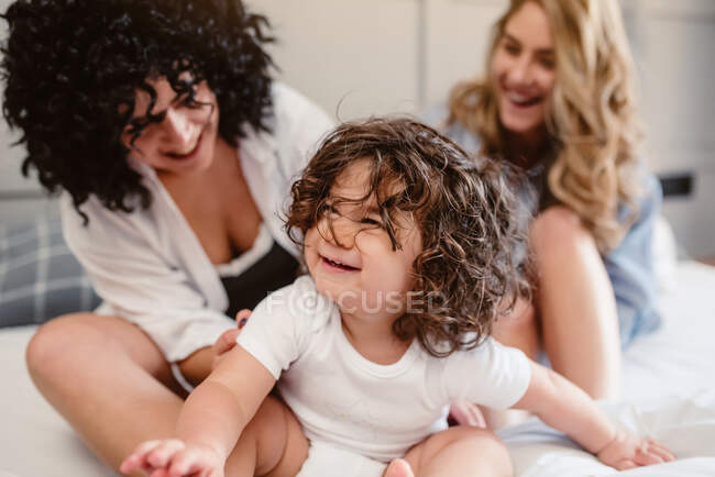 Positive crop same sex couple sitting on bed and stroking little curly haired girl in bedroom — Stock Photo