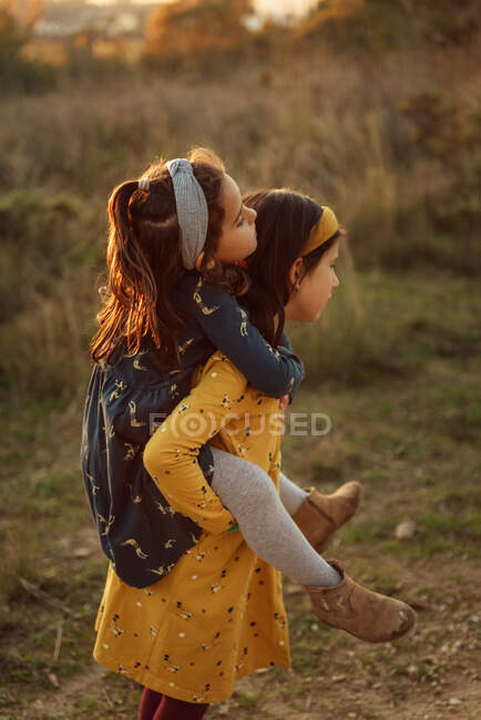Vista lateral de la amorosa niña preadolescente que lleva a la linda hermana pequeña en la espalda mientras pasan el día de verano juntos en el campo - foto de stock
