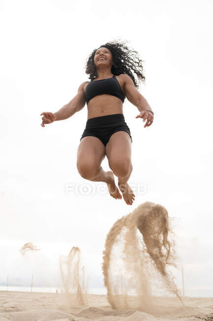 Bajo ángulo de alegre atleta asiática en el momento de saltar por encima de la orilla del mar de arena durante el entrenamiento de fitness en verano - foto de stock