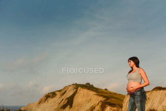 Vista lateral de pacífica joven hispana embarazada en jeans y parte superior de la cosecha revelando vientre de pie en la orilla del mar contra las montañas - foto de stock