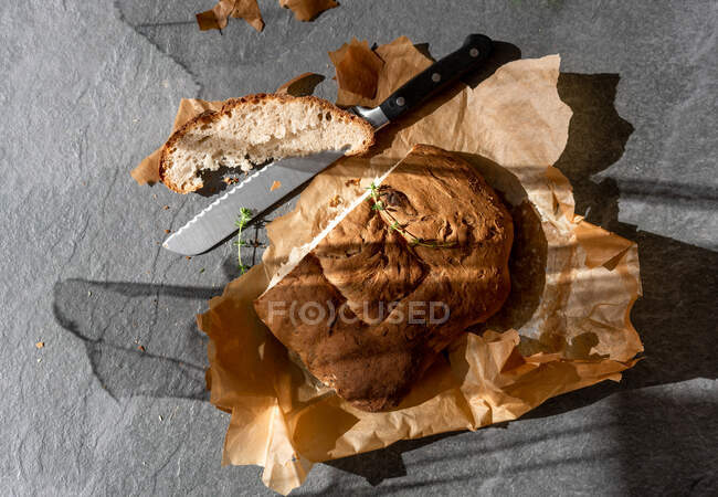 Vista superior de la deliciosa hogaza de pan de masa fermentada recién horneada con trozo en rodajas y cuchillo colocado sobre papel de hornear sobre fondo gris - foto de stock