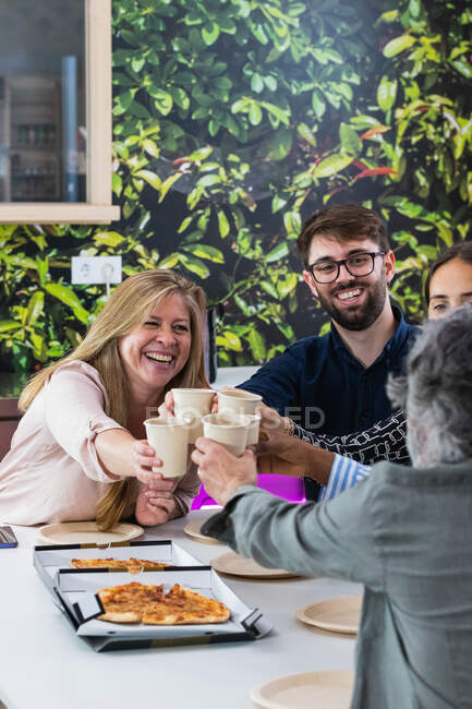 Gruppe zufriedener multiethnischer Mitarbeiter, die sich am Tisch versammeln und beim gemeinsamen Mittagessen in der Küche am Arbeitsplatz Tassen klappern — Stockfoto