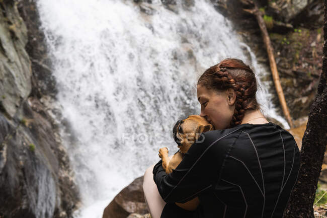 Розчарована жінка - туристка цілує французького бульдога біля водоспаду в лісі під час прогулянок у Піренеях. — стокове фото