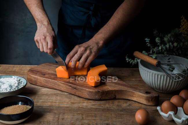 Невідомий чоловік готує в фартухах нарізаючи сирий гарбуз на дерев'яній обробній дошці біля борошна та хлібних крихт з насінням та яйцями під час приготування пирога — стокове фото
