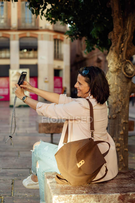 Вид сбоку веселый стильный взрослый латиноамериканец в модной повседневной одежде с сумкой, сидящей на скамейке и делающей селфи на смартфоне, отдыхая в одиночестве на городской улице — стоковое фото
