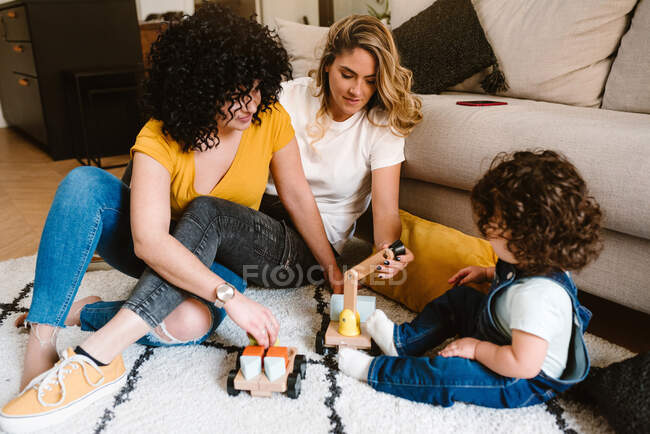 Conteúdo de corpo inteiro jovem casal lésbico em roupas casuais brincando com bebê bonito enquanto sentado no chão no apartamento moderno — Fotografia de Stock