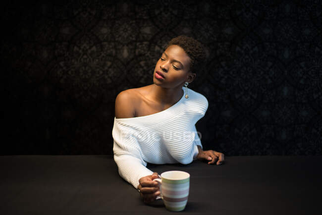 Безмятежная афроамериканка сидит за столом с кружкой освежающего напитка и смотрит в темную комнату — стоковое фото