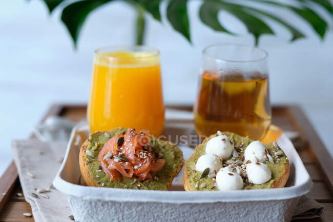Vasos de jugo y té de hierbas servidos en mesa de madera con una variedad de tostadas saludables de aguacate con queso y salmón durante el desayuno en la cafetería al aire libre - foto de stock