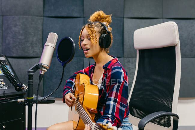 Молодая вокалистка в наушниках, играющая на акустической гитаре, поет с закрытыми глазами в микрофон в музыкальной студии — стоковое фото