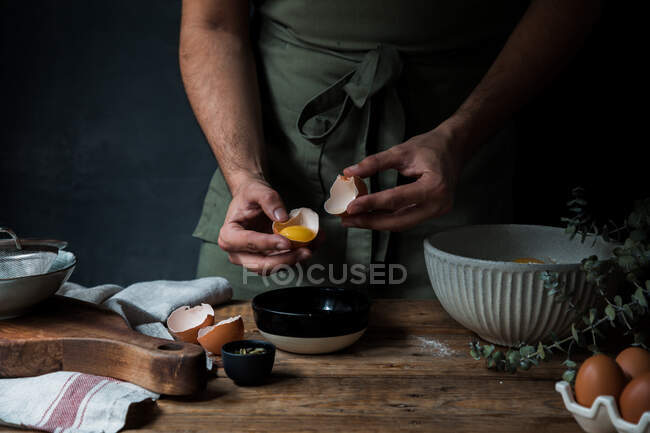 Unbekannter in Schürze bricht rohes Ei über Schüssel, während er Teig auf Holztisch in der Nähe von Geschirr zubereitet — Stockfoto