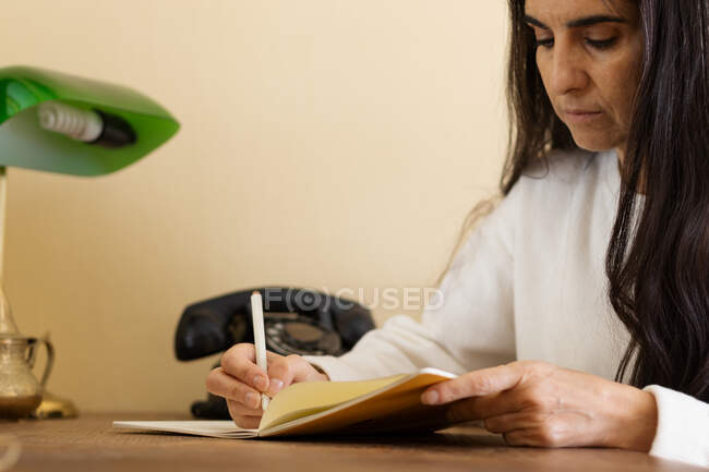 Mujer madura escribiendo en un cuaderno en un escritorio de madera con un teléfono viejo detrás. - foto de stock