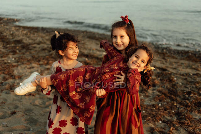 Cute little ethnic girls holding little sister on hands while having fun together on sandy beach near sea — Stock Photo