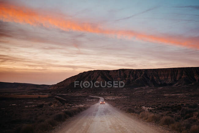 Veicolo veloce che guida su una strada di campagna arida vicino alle montagne al tramonto nel Parco Naturale Bardenas Reales in Navarra, Spagna — Foto stock