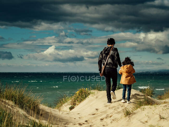 Обратный вид неузнаваемой женщины, держащейся за руку дочери, идущей вместе по холмистому песчаному побережью против волнистого океана и облачного неба в ветреную погоду — стоковое фото