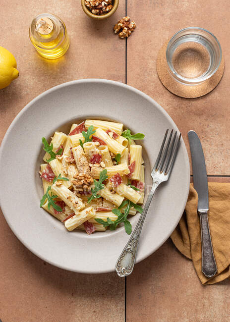 Dall'alto piatto di pasta con rucola, salame e noci su un tavolo circondato da olio, limone e posate — Foto stock