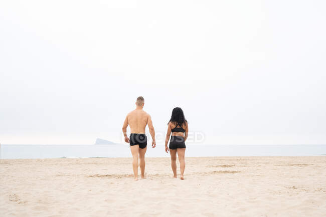 Обратный вид на неузнаваемую многорасовую спортсменку и спортсмена, прогуливающихся по песчаному побережью во время тренировки летом — стоковое фото