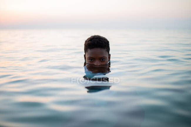 Mujer afroamericana con media cara en agua de mar mirando a la cámara en el fondo del cielo del atardecer - foto de stock
