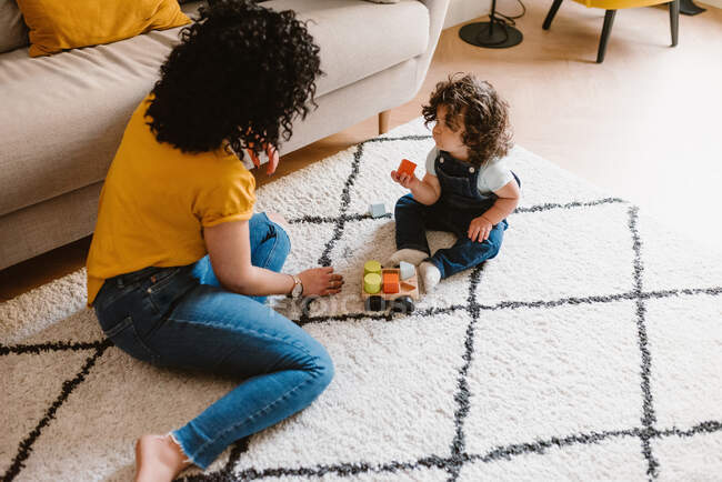 Madre en ropa casual jugando con un lindo niño pequeño mientras se sienta en el suelo juntos en la sala de estar - foto de stock