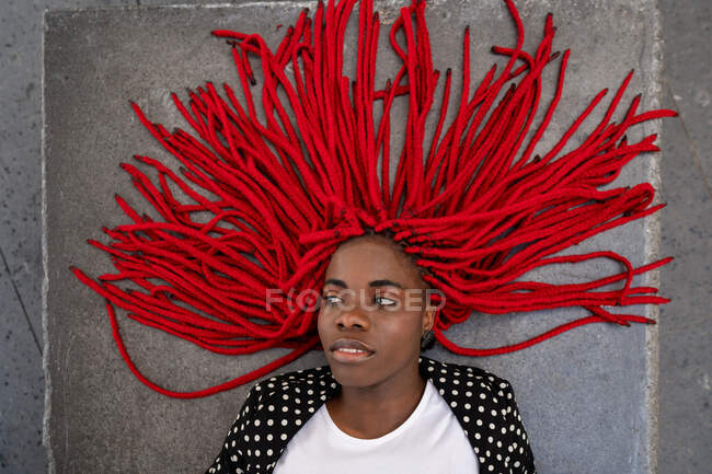 Draufsicht einer Afroamerikanerin mit roten Zöpfen, die auf dem Boden liegt und wegschaut — Stockfoto