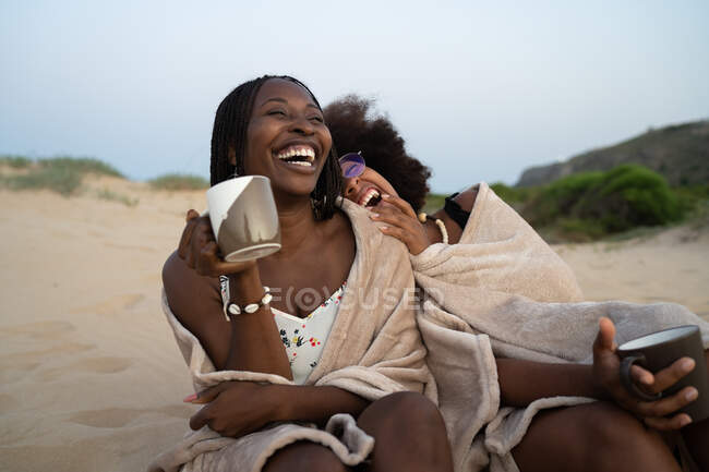 Feliz joven negro hembra mejores amigos con tazas de bebidas calientes sentado cerca envuelto en manta caliente y divertirse durante la noche de verano en la playa de arena - foto de stock