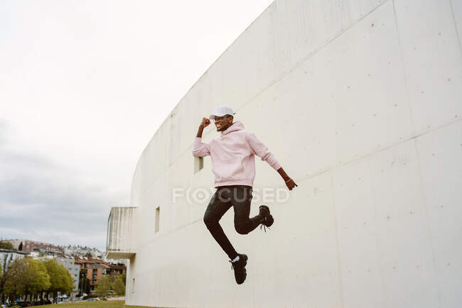 Low angle of full body of energetic African American male in casual wear laughing happily with fist up celebrating victory while jumping — Stock Photo