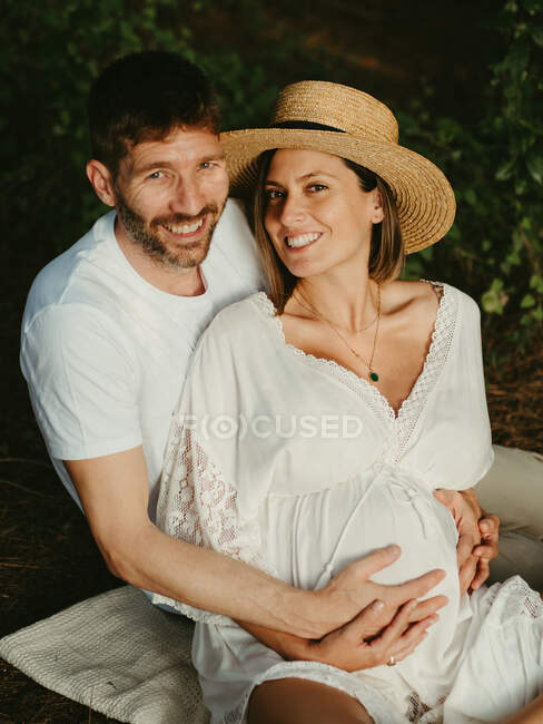 Smiling male hugging pregnant female from behind while sitting in countryside meadow looking at camera — Stock Photo