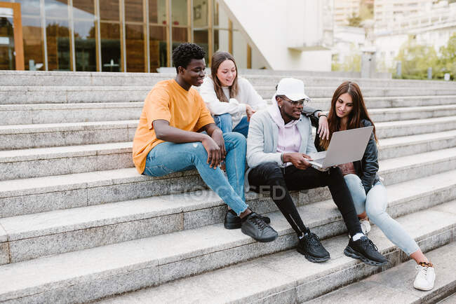 Різноманітні молоді друзі широко посміхаються під час перегляду відео в нетбуку, сидячи на бетонних сходах. — стокове фото