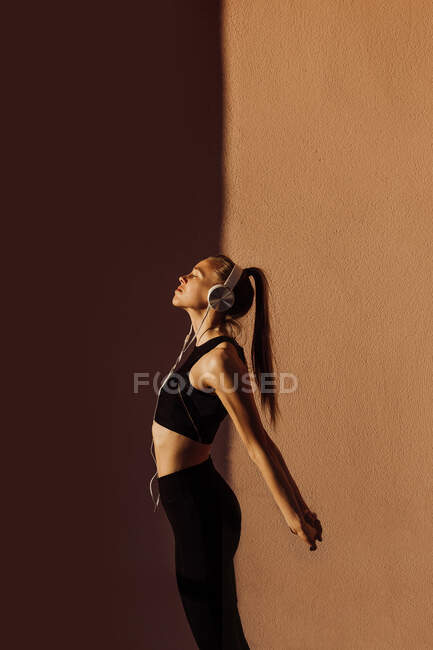 Vue latérale de la jeune femme caucasienne sportive écoutant de la musique et s'étirant au coucher du soleil, ombres et lumière sur le fond — Photo de stock