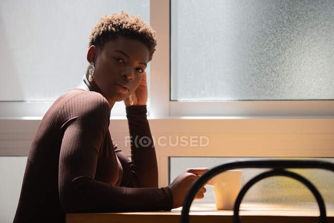 Side view of African American female with short hair drinking refreshing beverage from mug at home in morning looking at camera — Stock Photo