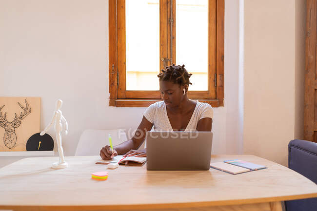 Freelancer afro-americano focado toma notas no caderno enquanto trabalha no projeto e se senta à mesa com laptop em casa — Fotografia de Stock
