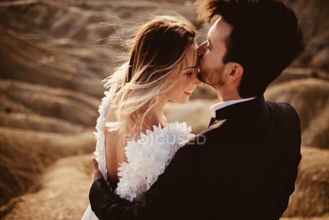 Hombre de traje abrazando y besando a mujer en la frente durante la celebración de la boda en el Parque Natural de Bardenas Reales en Navarra, España - foto de stock