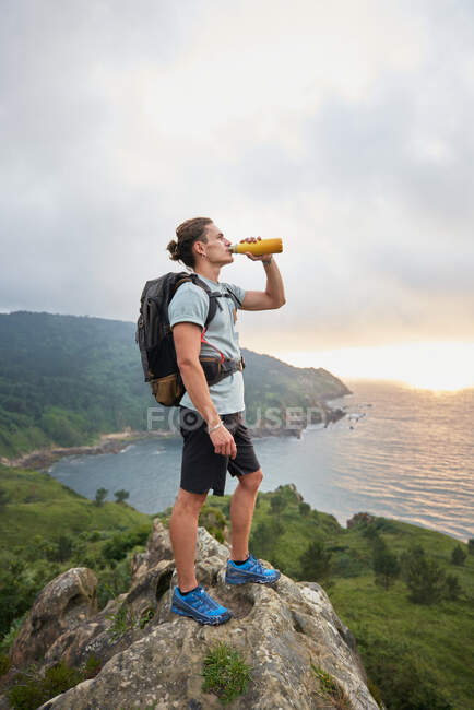 Vista lateral del caminante masculino de pie sobre piedra y agua potable sobre el fondo del cielo y el mar durante el trekking en verano - foto de stock