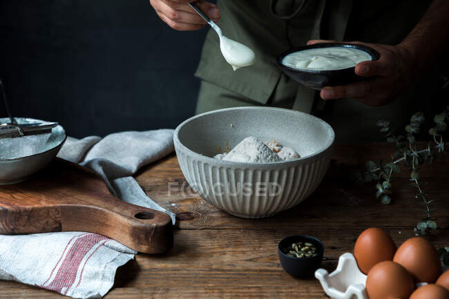 Unrecognizable person pouring yogurt into bowl with flour near eggs and seeds while preparing pastry on wooden table — Stock Photo