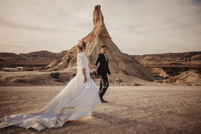 Mariée et marié heureux marchant main dans la main près de la montagne contre un ciel nuageux au coucher du soleil dans le parc naturel de Bardenas Reales en Navarre, Espagne — Photo de stock