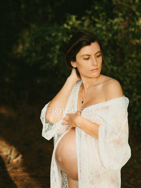Heitere schwangere Frau im Kleid, die den Bauch berührt, während sie im dunklen Wald steht und wegschaut — Stockfoto