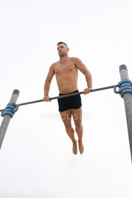 Низький кут м'язистого спортсмена з голим торсом робить підборіддя на горизонтальній планці під час тренувань на тлі сірого неба — стокове фото