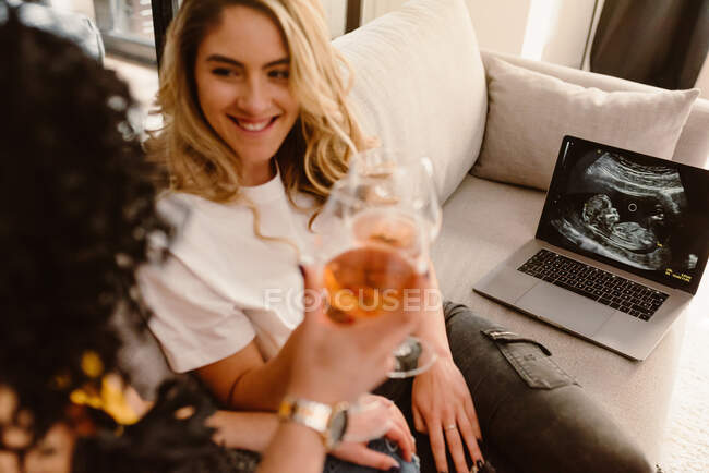 Обрезание улыбающейся лесбийской пары звон очков и смотреть друг на друга с нежностью во время празднования беременности и сидя на диване с ноутбуком, показывая ультразвуковое сканирование — стоковое фото