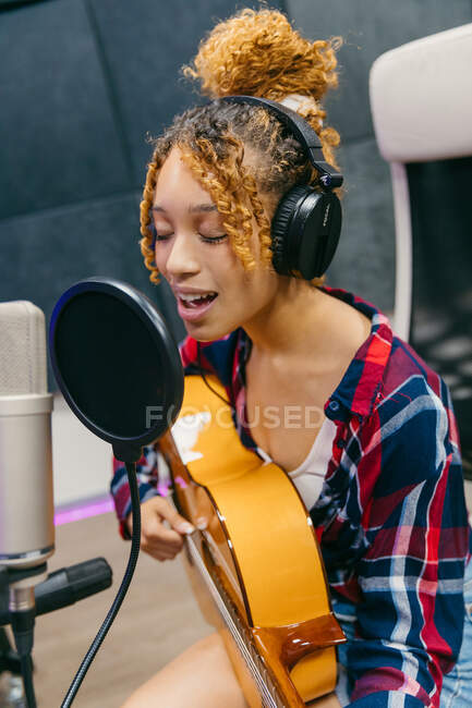 Young ethnic female vocalist in headphones playing acoustic guitar while singing with closed eyes into mic in music studio — Stock Photo