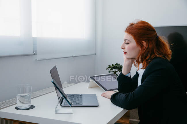 Side view of young female entrepreneur working on netbook at desk with tablet and smartphone at home — Stock Photo