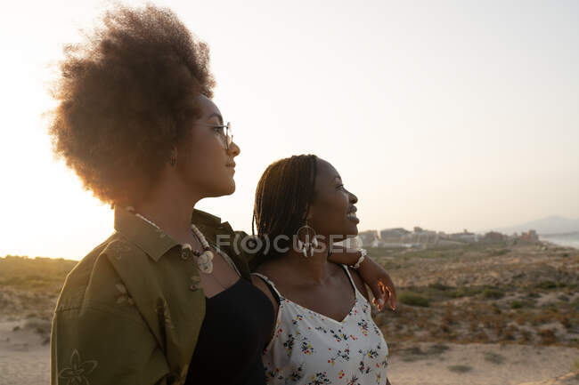 Счастливые молодые афро-американские подруги в стильных летних нарядах и аксессуарах обнимаются и смотрят в сторону, отдыхая вместе на берегу моря в летний вечер — стоковое фото