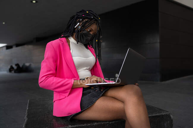 Side view full body of focused African American female in mask typing on netbook while working remotely outdoors — Stock Photo