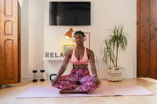 Mulher afro-americana relaxada sentada em Lotus posar e ouvir mediação em fones de ouvido enquanto faz ioga em casa — Fotografia de Stock