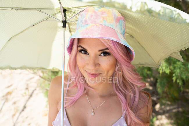 Angle élevé de femme joyeuse avec des cheveux roses se cachant sous le parapluie sur le bord de la mer le jour ensoleillé et regardant la caméra tout en profitant des vacances d'été — Photo de stock
