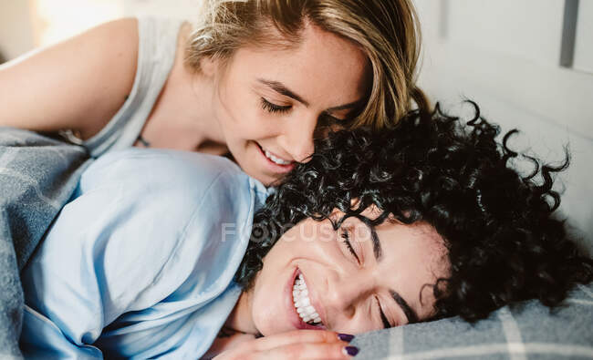 Cheerful crop girlfriend awakening curly haired lesbian partner lying in bed with closed eyes in morning — Stock Photo