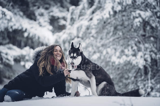 Бічний вид домашньої собаки бавиться з молодою леді на снігу між деревами в зимовий ліс. — стокове фото