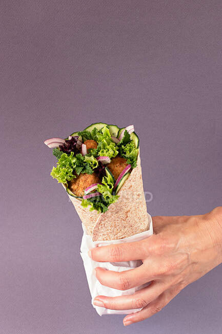 Cultivado irreconocible persona manos sosteniendo envoltura de falafel vegano sobre fondo púrpura colorido - foto de stock