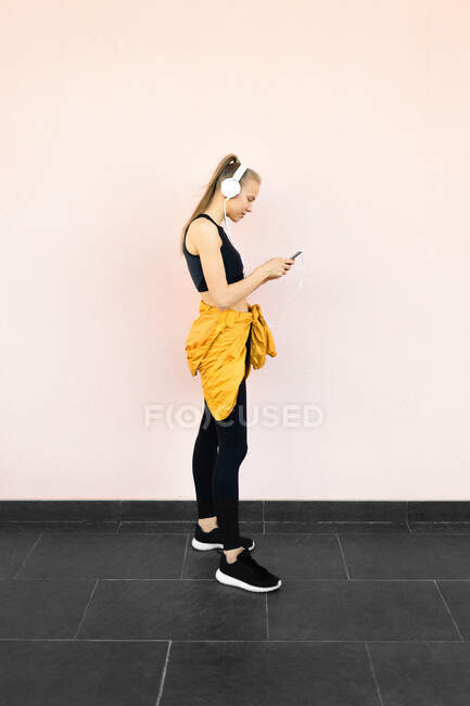 Jovem caucasiana vestindo fones de ouvido e roupas esportivas, ouvindo música no telefone, isolada em fundo brilhante — Fotografia de Stock